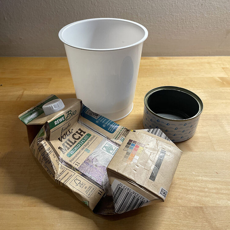 Eine zerknüllte Milchpackung neben einem weißen Kunststoffbecher und einer geöffneten, leeren Konservendose auf einer hellen Holzfläche vor einer hellen Wand.