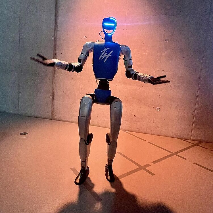 Ein blau-weiß humanoider Roboter mit leuchtend blauem Visier steht in einem hell erleuchteten Ausstellungsraum vor einer Betonwand, die Arme seitlich ausgestreckt; sein Schatten und markierte Bodenkreuze sind auf dem Fußboden sichtbar.