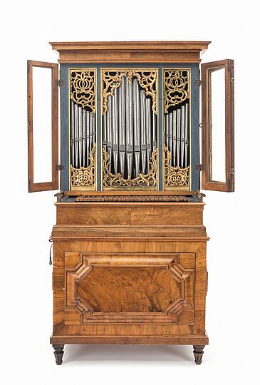 Das Bild zeigt ein das Lamprecht-Orgelpositiv.  Ein Orgel-Positiv ist eine Orgel, die im Format einem Schrank ähnelt. Bei diesem Exemplar ist die Klaviatur direkt vor den Pfeifen angeordnet. Von diesen sehen wir drei Werke, also Pfeifengruppierungen umgeben von geschwungenen hölzernen Verzierungen.