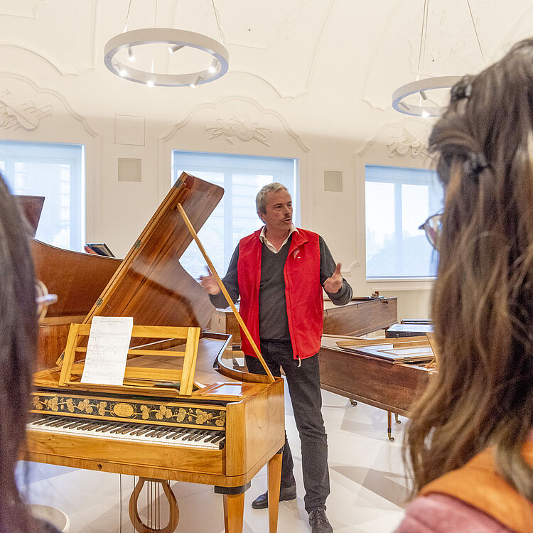 Ein Museumsausstellungsraum mit historischen Tasteninstrumenten: im Vordergrund ein aufgeklappter, hellholzfarbener Flügel mit verziertem Notenpult und Notenblatt, im Hintergrund eine stehende Person mit roter Weste (Gesicht unkenntlich) die offenbar erklärt; zwei Besucherinnen im Vordergrund von hinten gesehen; helle Innenraumbeleuchtung, dekorative Stuckdecke und große Fenster.