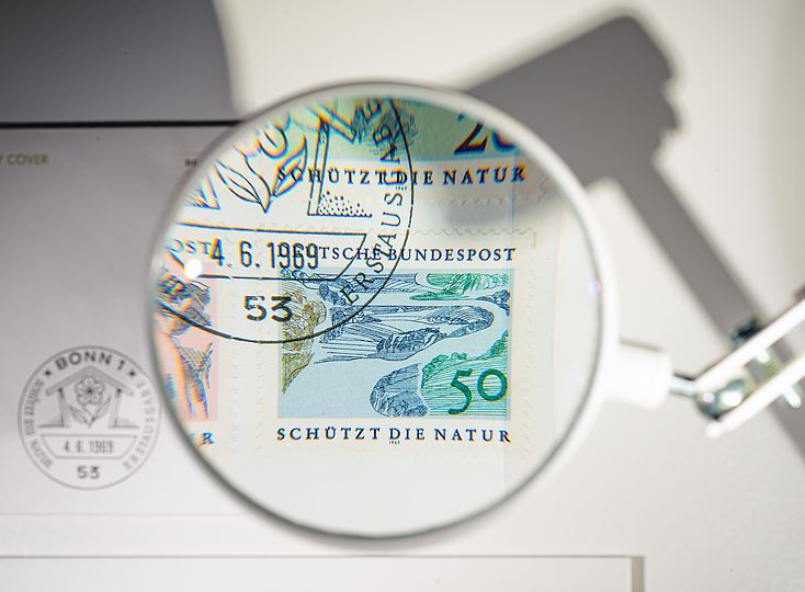 Kleine Lupe vergrößert eine der vier Briefmarken und einen Teil des Stempels. Die Briefmarke zeigt einen Fluss, der sich durch die Natur schlängelt. Zu lesen ist "Schützt die Natur".