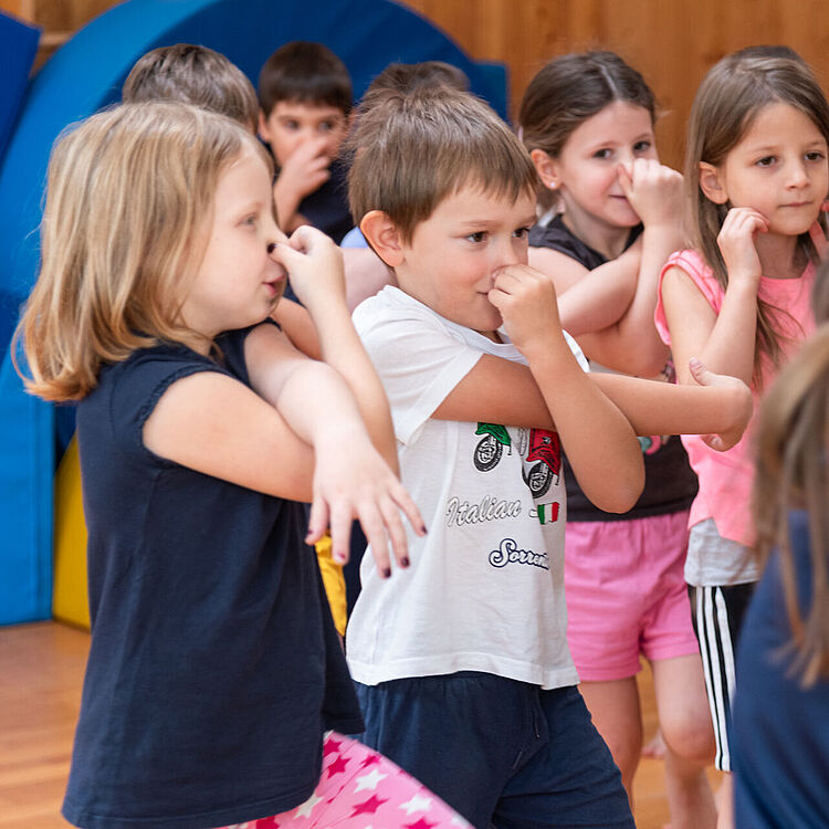 Eine Gruppe von Grundschulkindern in Turnhalle macht Atemübungen: mehrere Mädchen und ein Junge stehen dicht beieinander, halten sich die Nasen oder pressen Finger an die Nasenwurzel, tragen bunte Freizeitkleidung (T-Shirt, Shorts), im Hintergrund blaue Weichbodenmatten und Holzverkleidung.