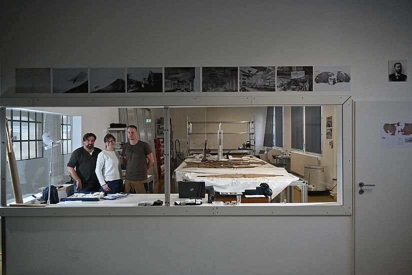 Blick in eine Werkstatt auf der auf einem Tisch der Gleiter liegt, daneben steht das Restauratoren-Team. 