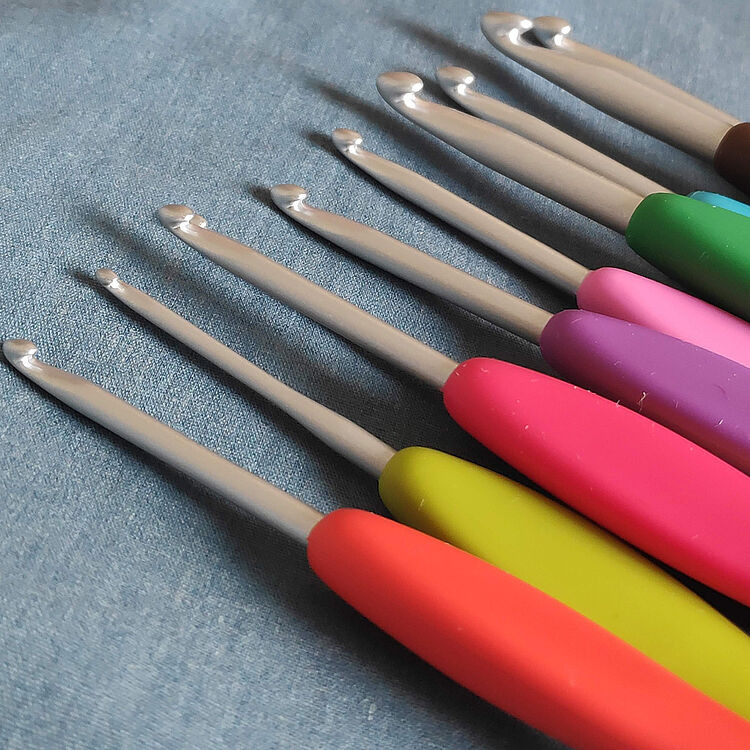 Nahaufnahme mehrerer Häkelnadeln unterschiedlicher Größe mit farbigen Griffen in Rot, Gelb, Pink, Lila, Grün und Blau. Die Metallspitzen liegen fächerförmig auf einem hellblauen Stoff und werfen weiche Schatten. Die Aufnahme betont die verschiedenen Größen und Farben der Werkzeuge.