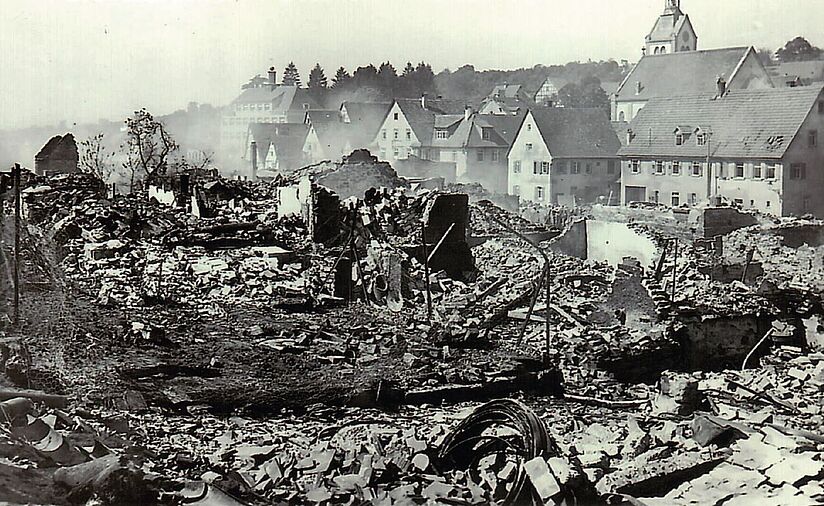 Die Schwarzweiß-Fotografie zeigt einen zum Teil verbrannten Ort. Aus den Ruinen der Häuser steigt noch Rauch auf.