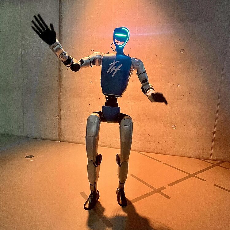 Ein menschenförmiger Roboter mit blauem Oberkörper und leuchtender, cyanfarbener Gesichtskontur steht in einem warm beleuchteten Raum vor einer Betonwand, hebt die linke Hand zum Winken; starker Schatten des Roboters liegt auf dem Boden.