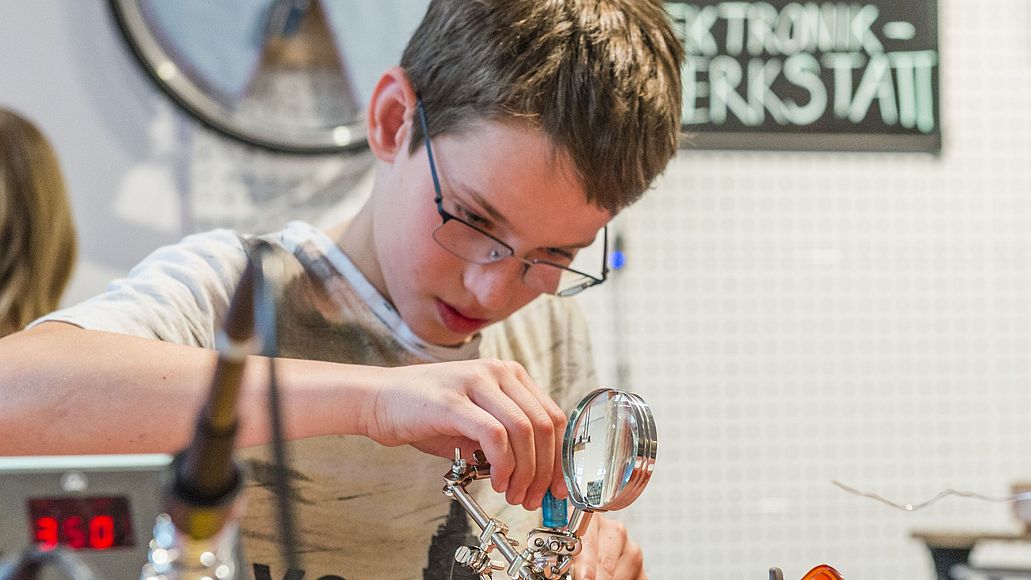 Elektronik-Werkstatt Junge lötet an einem Tisch eine kleine Figur.