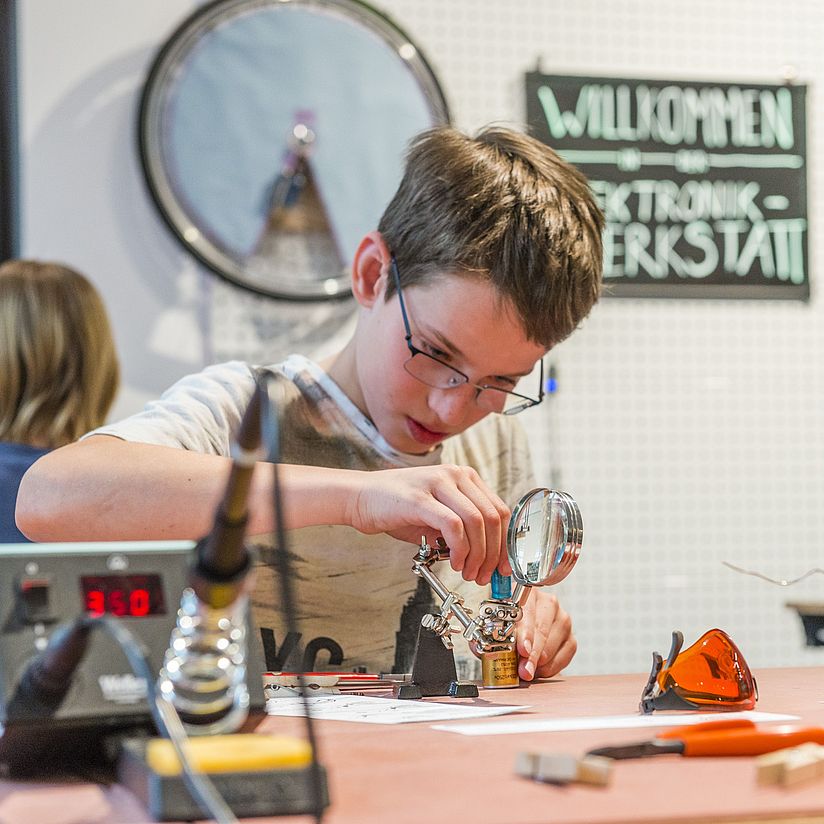 Elektronik-Werkstatt Junge lötet an einem Tisch eine kleine Figur.