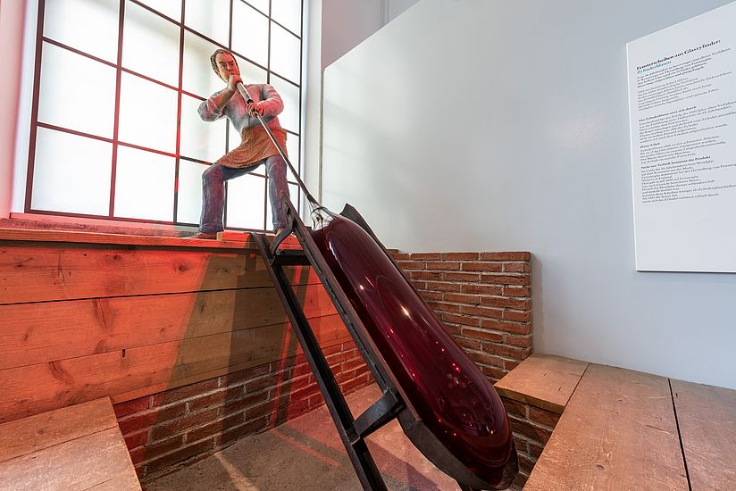 Life-size figure with a blowpipe. It shows how container glass is formed manually from molten glass with a blowpipe.