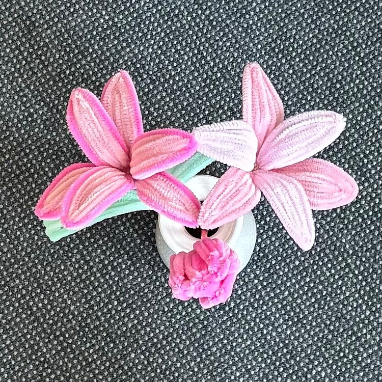 Nahaufnahme einer kleinen DIY-Blumenanordnung aus rosa und hellrosa Blüten und grünen Stiehlen aus Pfeifenputzern, platziert in einer kleinen weißen Vase auf dunkelgrauem Untergrund.