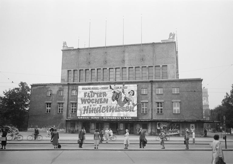 Schwarzweiß Fotografie des Forums der Zukunft mit Menschen im Vordergrund. Auf einem Plakat wird ein Film angekündigt.