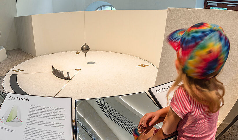 Ein Mädchen  beobachtet aufmerksam das Foucault’sches Pendel, das gleichmäßig über einer kreisrunden Scheibe schwingt.