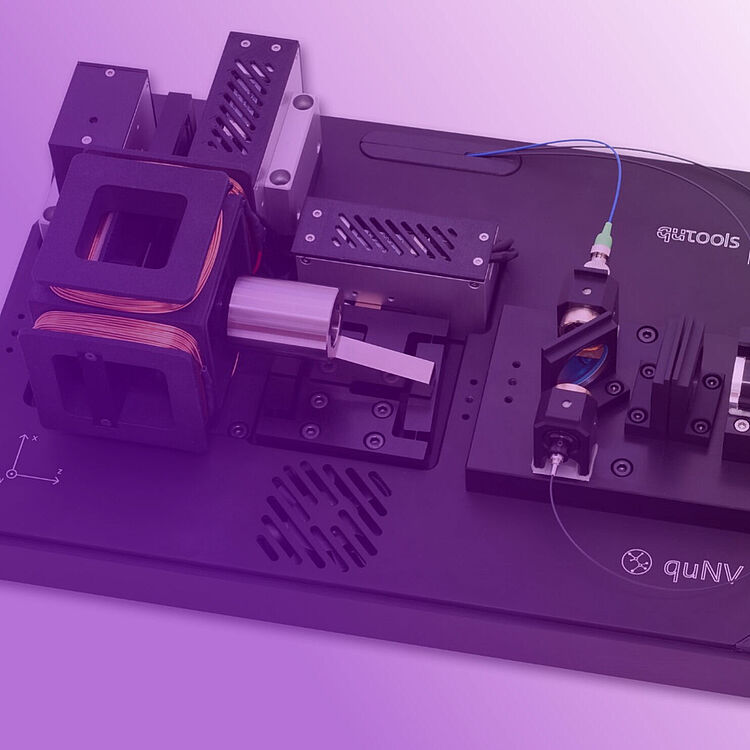 Ein schwarzes Laborprüfbrett mit elektronischen Komponenten: links zwei quadratische Spulenkerne mit Kupferwicklungen und einem zylindrischen Motor, in der Mitte Metallgehäuse und Montageplatte mit Schrauben, rechts Faseroptikstecker, Glasfaserleitung und optische Halterungen auf einer beschrifteten Basisplatte mit Firmenlogo "guTools / quNV".