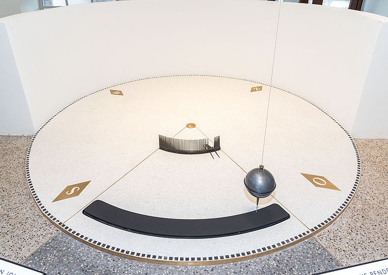 Nahaufnahme des Foucault’schen Pendels im Museumsturm. Kreisrunde Scheibe von 3 m Durchmesser über ihr die schwingende Bleikugel.