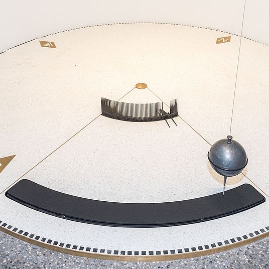 Nahaufnahme des Foucault’schen Pendels im Museumsturm. Kreisrunde Scheibe von 3 m Durchmesser über ihr die schwingende Bleikugel.