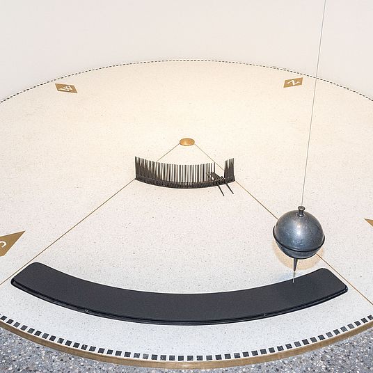 Nahaufnahme des Foucault’schen Pendels im Museumsturm. Kreisrunde Scheibe von 3 m Durchmesser über ihr die schwingende Bleikugel.