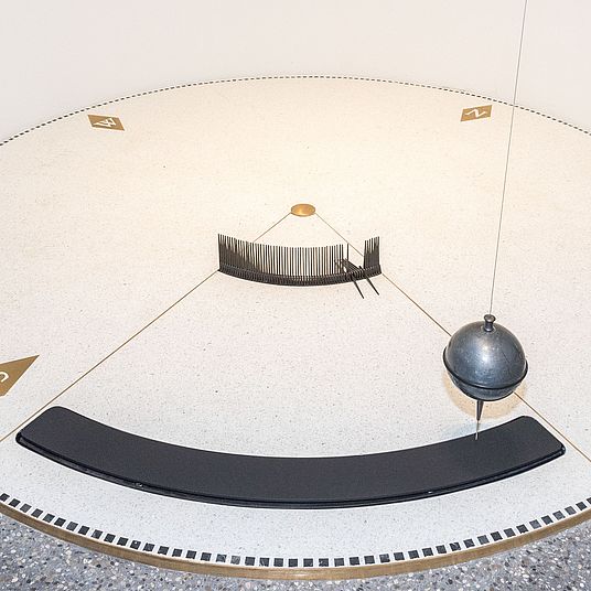 Nahaufnahme des Foucault’schen Pendels im Museumsturm. Kreisrunde Scheibe von 3 m Durchmesser über ihr die schwingende Bleikugel.