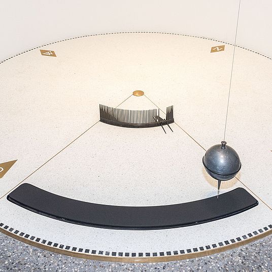 Nahaufnahme des Foucault’schen Pendels im Museumsturm. Kreisrunde Scheibe von 3 m Durchmesser über ihr die schwingende Bleikugel.