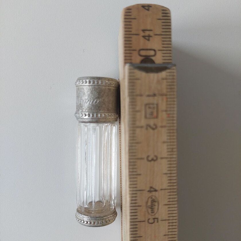 Miniflakon (7,5 ml, ca. 5 cm hoch) des Parfums Dior Diorissimo aus gerilltem Glas mit metallenem Stöpsel neben einem hölzernen Meterstab