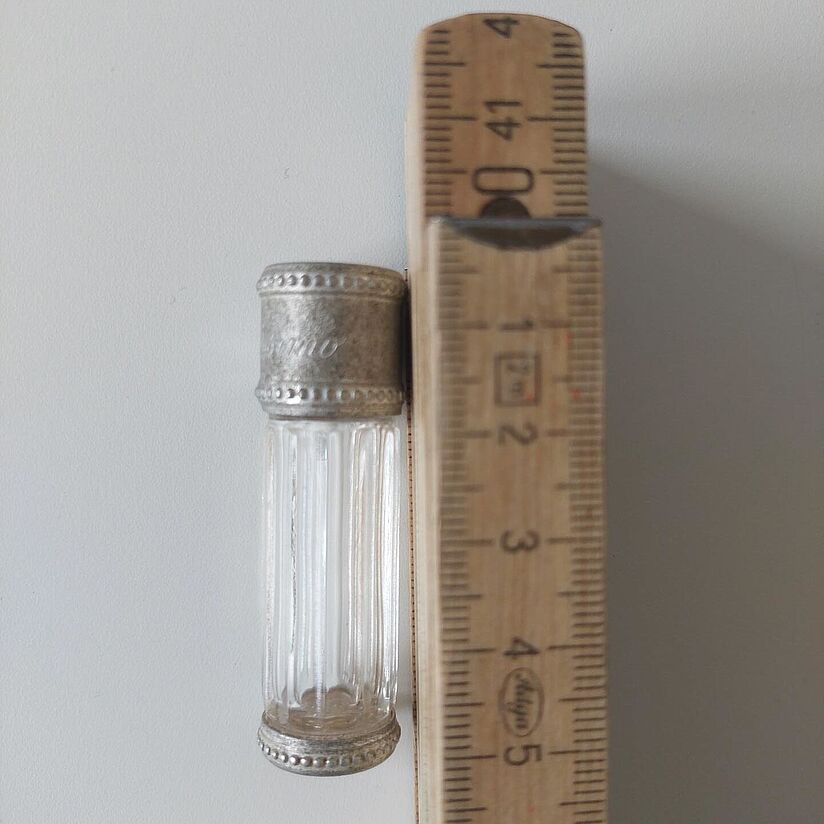 Perfumflakon Dior Diorissimo Miniflakon (7,5 ml, ca. 5 cm hoch) des Parfums Dior Diorissimo aus gerilltem Glas mit metallenem Stöpsel neben einem hölzernen Meterstab