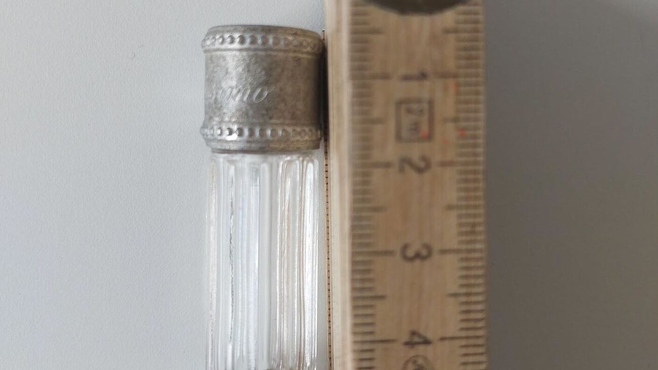 Miniflakon (7,5 ml, ca. 5 cm hoch) des Parfums Dior Diorissimo aus gerilltem Glas mit metallenem Stöpsel neben einem hölzernen Meterstab