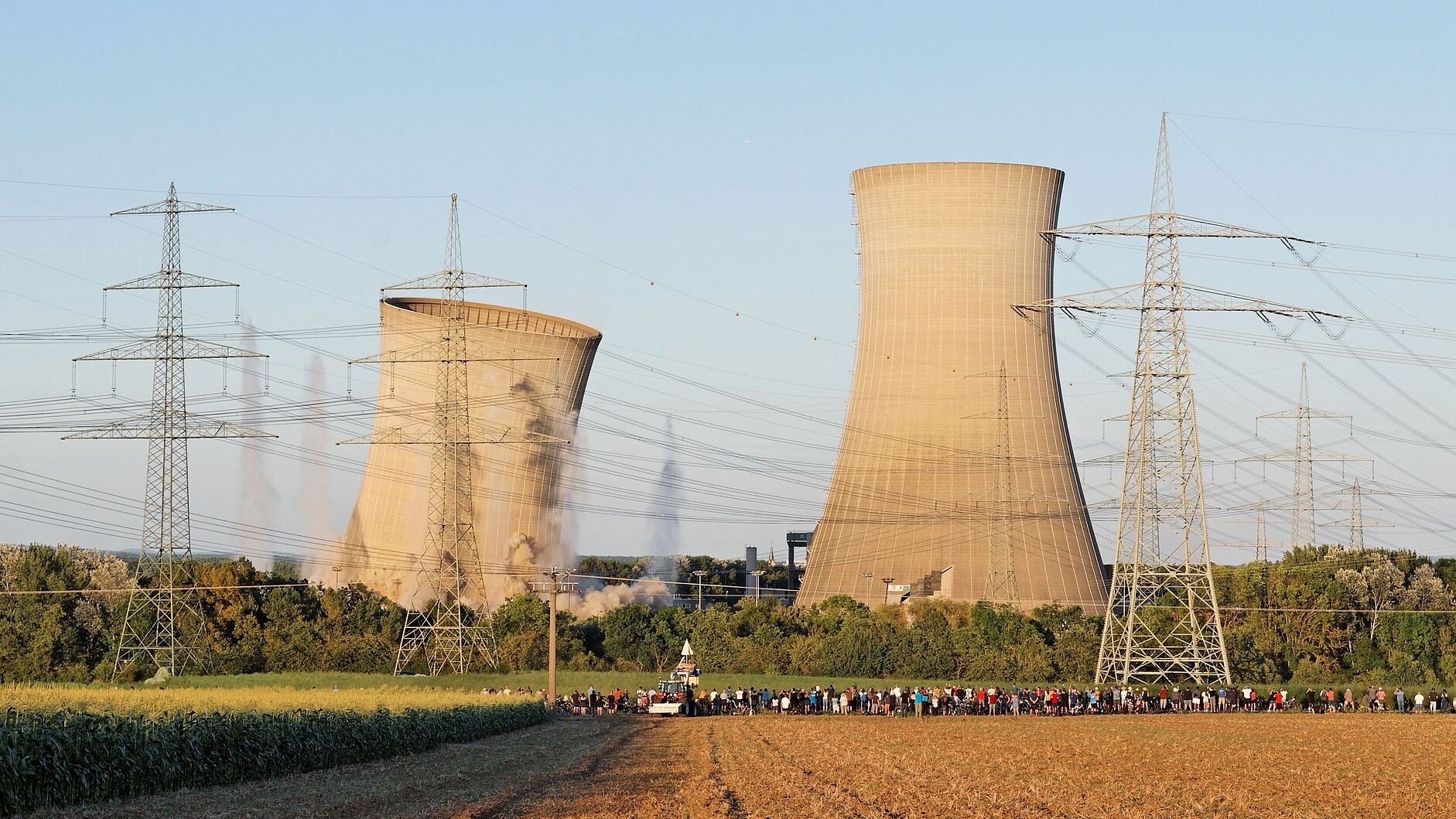 Im Vordergrund sind ein Feld, Strommasten und in einiger Entfernung eine Ansammlung von Menschen. Im Hintergrund: zwei Kühltürme eines Atomkraftwerks, der linke bricht nach rechts ein, am Boden ist Staub von der Sprengung zu sehen.