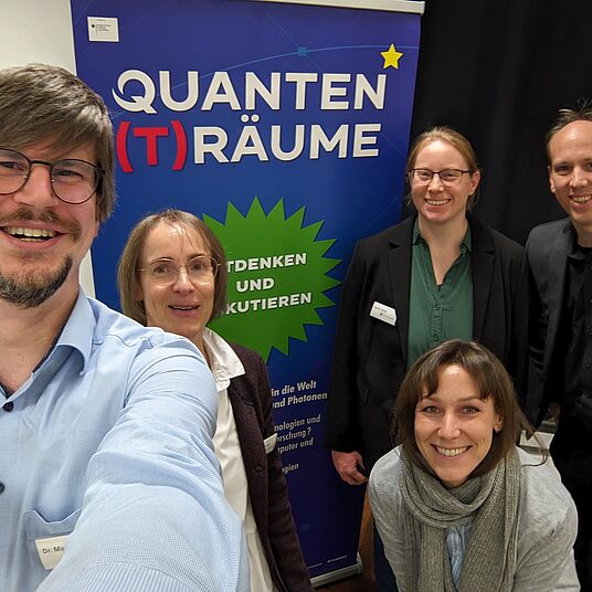5 Menschen lachen begeistert in die Kamera. Im Hintergrund ist das Roll-Up mit dem Schriftzug Quanten(t)räume.