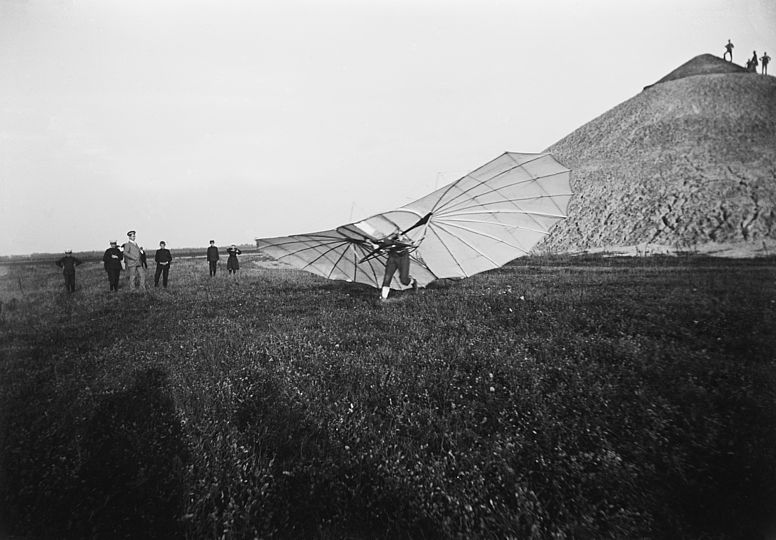 Lilenthal steht auf der Wiese, mit beiden Füßen noch in Bewegung, circa 10 Zuschauer stehen bei ihm im Hintergrund der Fliegeberg.