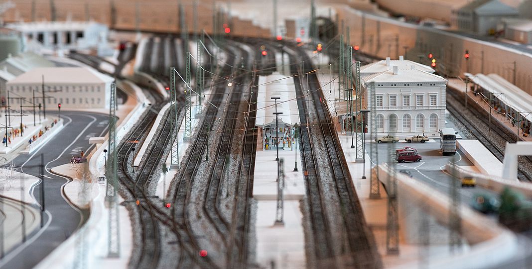 Detail der Modellbahn. Zu sehen ist der Bahnhof mit Parkplatz, zwei überdachte Bahnsteige und Gleise.