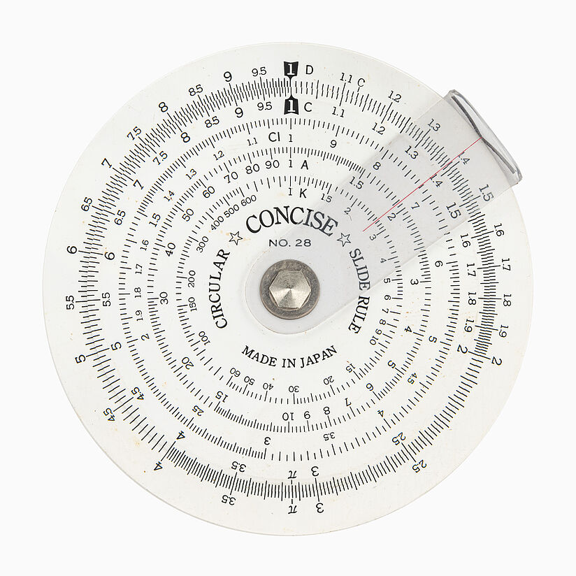 Weiße, runde Scheibe mit in kreisrichtung verschiebbarer Plastik Lasche (roter Strich radial). Mehrere Zeilen mit Skalen und Zahlen kreisförmig angeordnet.