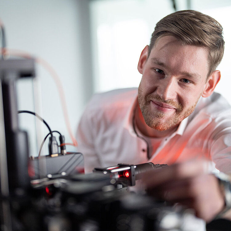 Ein lächelnder junger Mann in weißem Hemd, der in einem Laborumfeld an einer komplexen elektronischen Versuchsanordnung mit Kabeln, Sensoren und roten Laser- oder LED-Punkten arbeitet und ein Bauteil justiert.