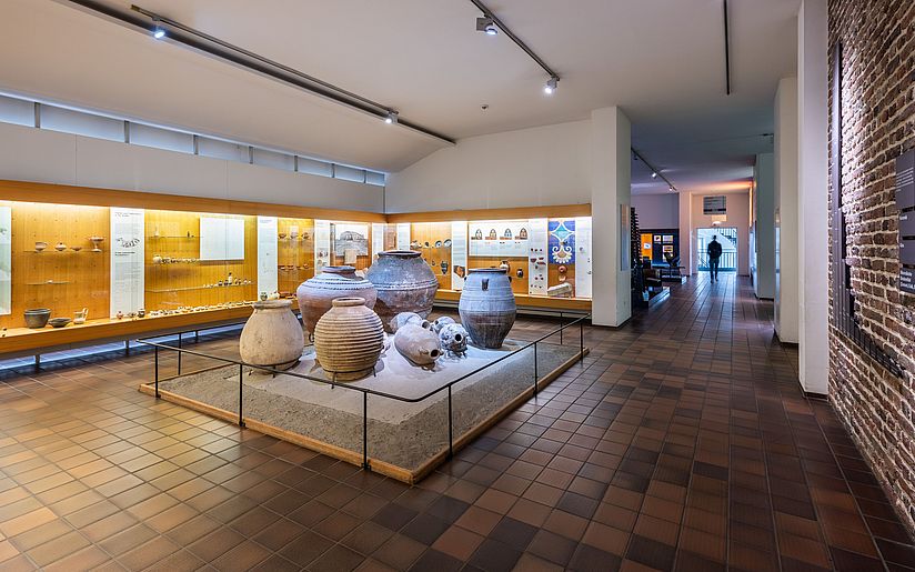 Raumansicht Ausstellung Keramik. An den Wänden befinden sich große Vitrinen mit Obejekten aus Keramik. In der Mitte des Raums befinden sich große Vorratsgefäße.