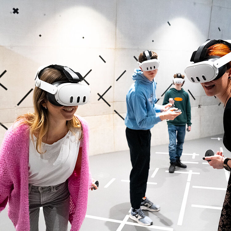 Eine Gruppe junger Menschen in einem hellen, minimalistischen Raum trägt weiße Virtual-Reality-Headsets und hält Controller; vorne links eine Frau mit rosa Strickjacke, vorne rechts eine Frau mit roten Haaren und schwarzem Oberteil, im Hintergrund zwei jüngere Personen in blauen und grünen Pullovern; Wand mit schwarzen Kreuz- und Strichmarkierungen, Boden mit weißen Markierungen.