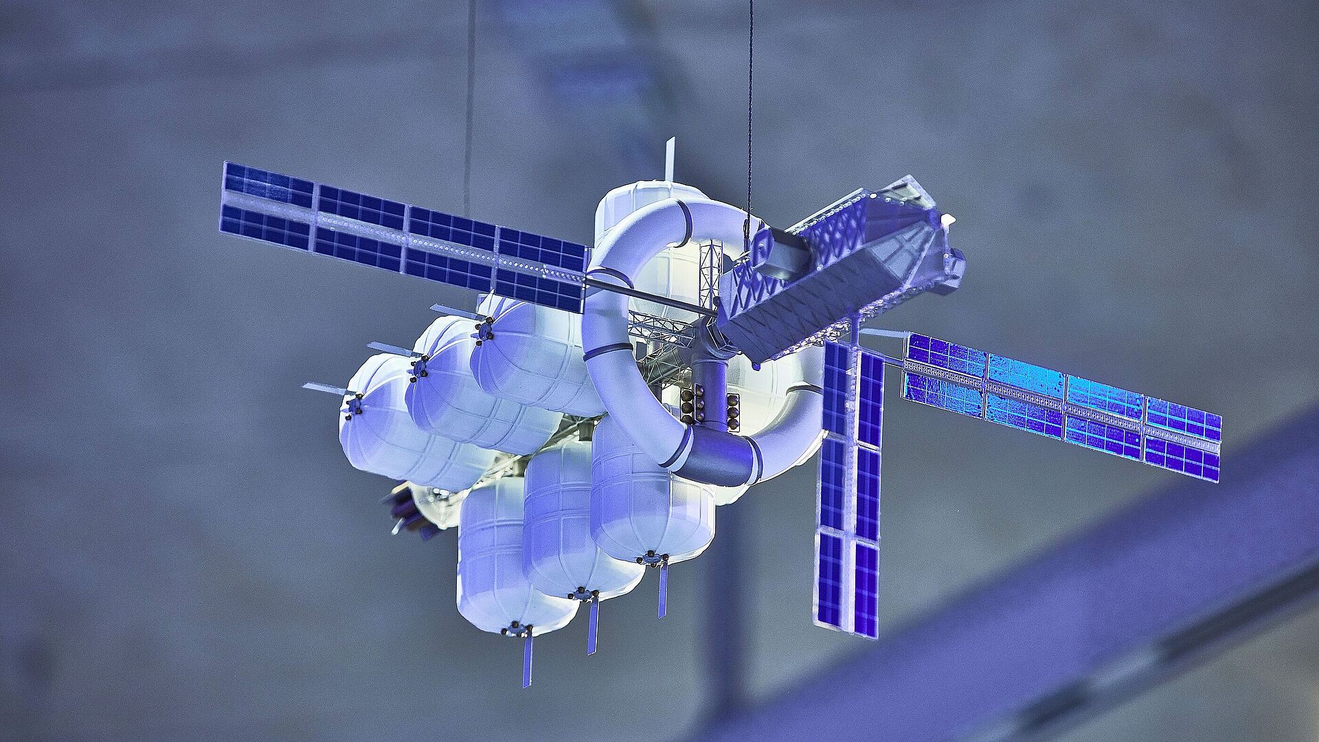 Detailliertes Modell einer futuristischen Raumstation mit zylindrischen Druckmodulen, ringförmiger Verbindungsstruktur und vier blauen Solarpanelen, an dünnen Drähten hängend vor unscharfem graublauem Hintergrund.