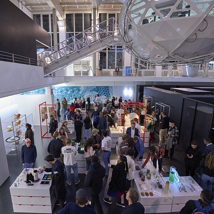 Eine große, moderne Ausstellungshalle aus der Vogelperspektive mit zahlreichen Besucherinnen und Besuchern, zentralen Ausstellungstischen und Vitrinen, roten Rahmenstrukturen und einer Galerieebene mit Treppe im Hintergrund.