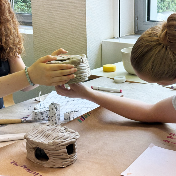 Zwei Personen arbeiten gemeinsam an kleinen Tonmodellen. Eine Person hält ein rundes Hausmodell aus Ton, während die andere Person eine Hand ausstreckt, um zu helfen. Auf dem Tisch liegen Werkzeuge, Tonreste und ein weiteres Modell.