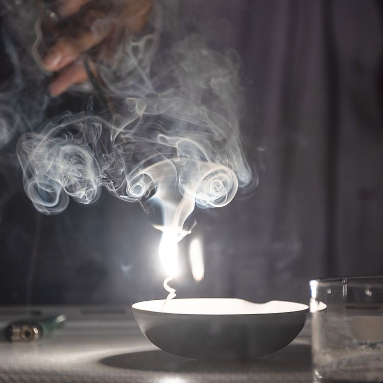 Eine Mitarbeiterin der Ausstellung Chemie zündet im Labor Salze an und eine weiße Flamme und Rauch sticht empor.
