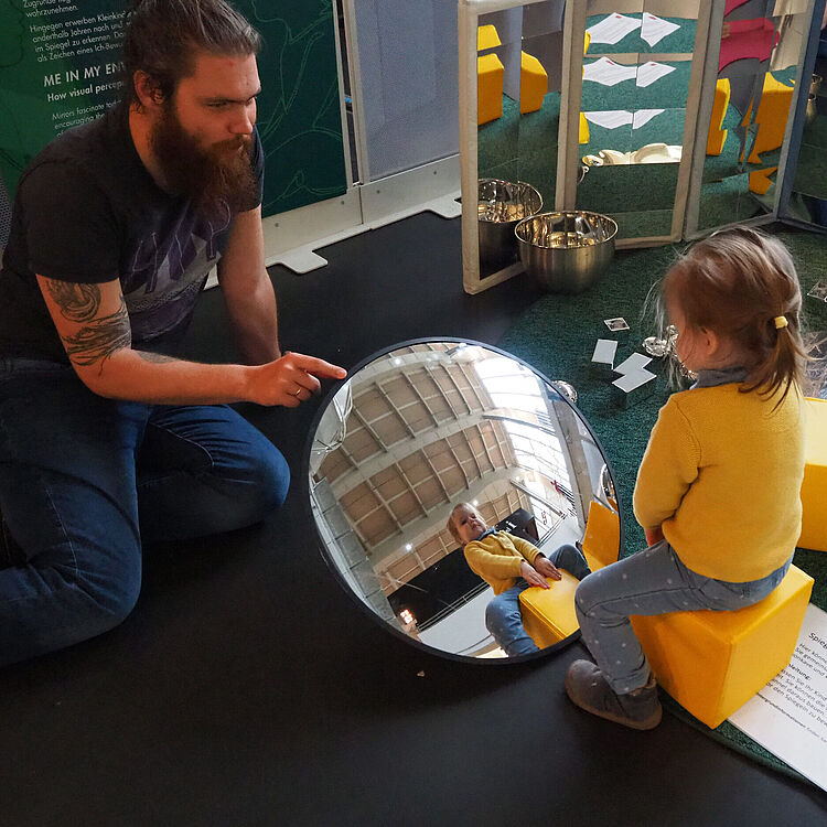 Ein Mann und ein kleines Kind sitzen sich gegenüber und betrachten gemeinsam ihr Spiegelbild in einem großen, runden, gewölbten Spiegel auf dem Boden. Das Kind sitzt auf einem gelben Würfelhocker, umgeben von weiteren Materialien in einem interaktiven Museumsbereich.
