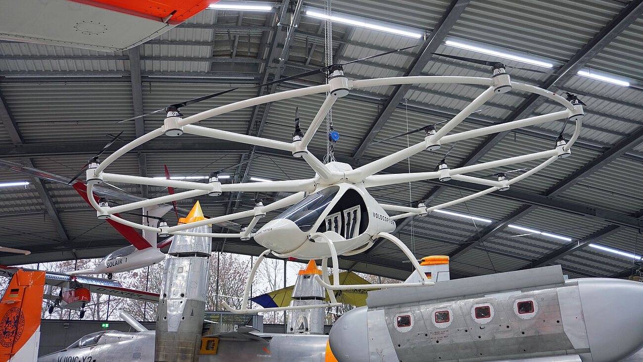 Fotografie von Halle in Richtung Decke. Zentral ist weißes Fluggeräte mit Kabine und 18 auf zwei Ringen angeordneten nach oben gerichteten Rotoren.