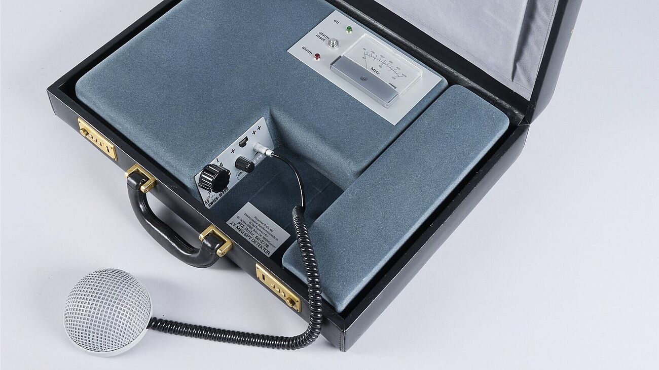 Schwarze, geöffneter Lederkoffer. Darin eingelegt: mit Samt umzogener Apparat mit MHz Anzeige, drei Lämpchen und einem Mikrofonanschluss mit Reglern.
