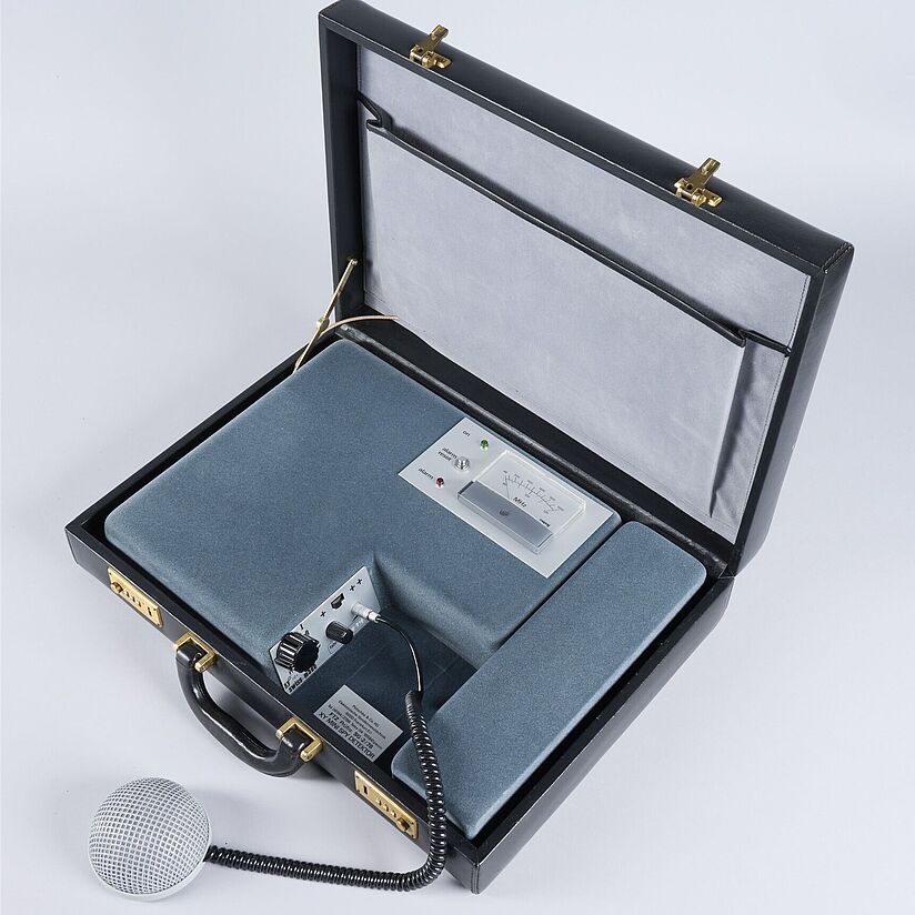 Schwarze, geöffneter Lederkoffer. Darin eingelegt: mit Samt umzogener Apparat mit MHz Anzeige, drei Lämpchen und einem Mikrofonanschluss mit Reglern.