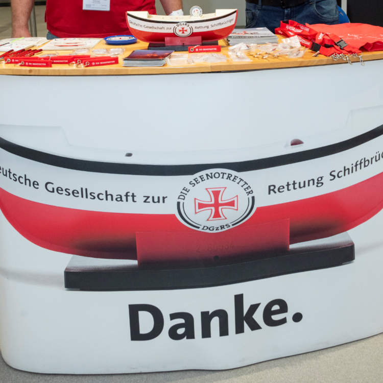 Messestand-Tresen mit Aufdruck einer roten Rettungsboje und dem Schriftzug "Deutsche Gesellschaft zur Rettung Schiffbrüchiger" sowie großem "Danke." auf der Vorderseite; auf der Holzplatte oben Werbematerialien, Schlüsselbänder und eine kleine Modell-Rettungsbootschale.