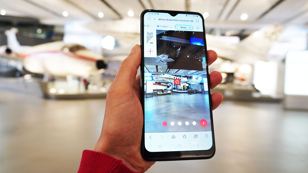 Handy in Hand, der Bildschirm zeigt das gleiche Bild wie vor dem Handy: einen Ausschnitt der modernen Luftfahrt Ausstellung.