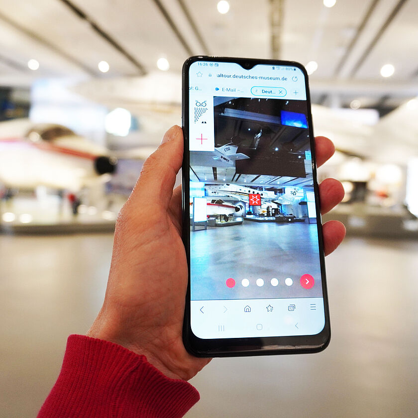 Handy in Hand, der Bildschirm zeigt das gleiche Bild wie vor dem Handy: einen Ausschnitt der modernen Luftfahrt Ausstellung.