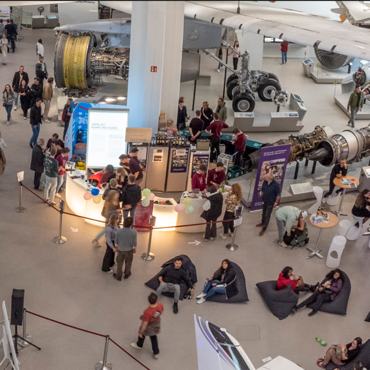 Vogelperspektive auf eine belebte Ausstellungshalle eines Luftfahrtmuseums: Besucher schlendern zwischen großen Flugzeugtriebwerken, freistehenden Exponaten und Informationsständen, in der Mitte ein halbkreisförmiger, beleuchteter Infopoint mit Ballons und Warteschlange, rechts Gruppen an Stehtischen und Sitzsäcken, oberhalb Flugzeugflügel und Fahrwerk sichtbar.