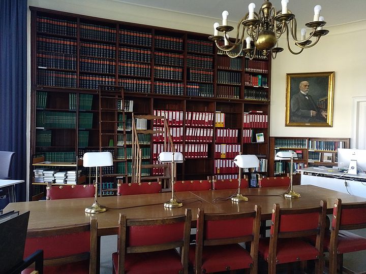 Alter Lesesaal mit einem großen Regal mit Aktenordnern. Ein langer Holztisch mit vier Stühlen steht davor. Auf dem Tisch stehen vier Lampen. Rechts über dem Tisch hängt ein großer Kronleuchter. An der  rechten Wand ein Portrait von Max Planck.