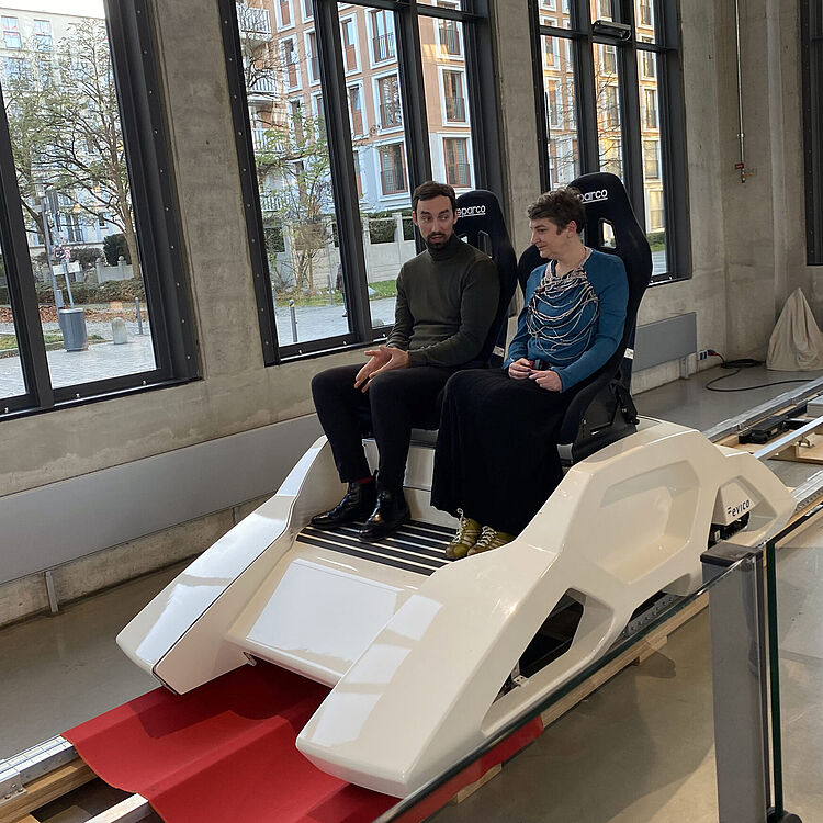 Zwei Personen testen den Supra-Gleiter, einen weißen Prototyp mit zwei Schalensitzen auf Schiene.