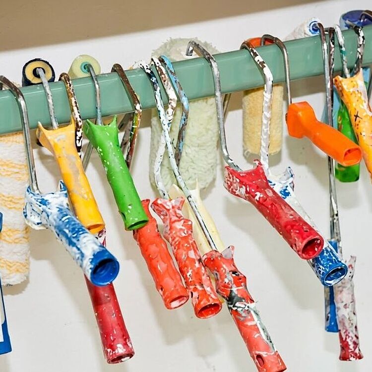 Bunte Rollenhalter und Farbpinselgriffe in Grün, Blau, Gelb, Orange und Rot mit Farbspritzern hängen an einer grünen Wandhalterung; dahinter mehrere benutzte Farbrollen mit Farbresten.