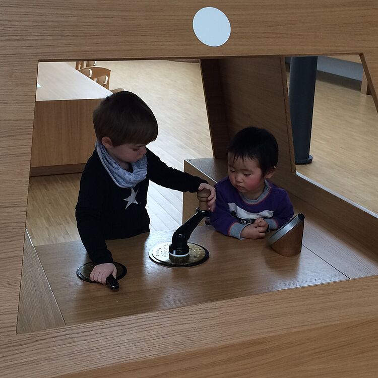 Zwei Kinder mit verpixelten Gesichtern sitzen in einem hellen, hölzernen Spielhäuschen und bedienen gemeinsam ein schwarzes Handrad sowie eine runde Metallplatte auf einer Holzoberfläche; das Häuschen hat große rechteckige Öffnungen und einen weißen Kreisaufkleber oben.