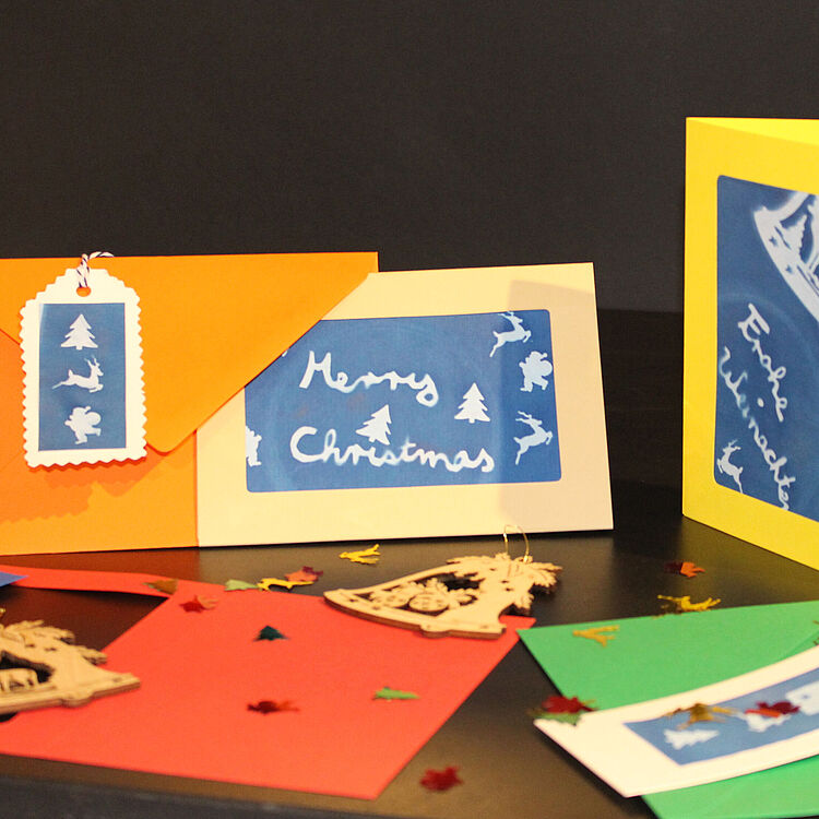 Ein Set aus handgemachten Weihnachtskarten und Umschlägen, das auf einer dunklen Fläche präsentiert wird. Die Karten zeigen blaue Ausschnitte mit weißen weihnachtlichen Motiven wie Bäumen, Rentieren und Schneeflocken, mit Aufschriften wie „Merry Christmas“ und „Frohe Weihnachten“. Die Umschläge sind leuchtend in Orange, Gelb und Grün gehalten. Dekorative Weihnachtsanhänger aus Holz und sternförmiges Konfetti sind um die Karten verstreut.